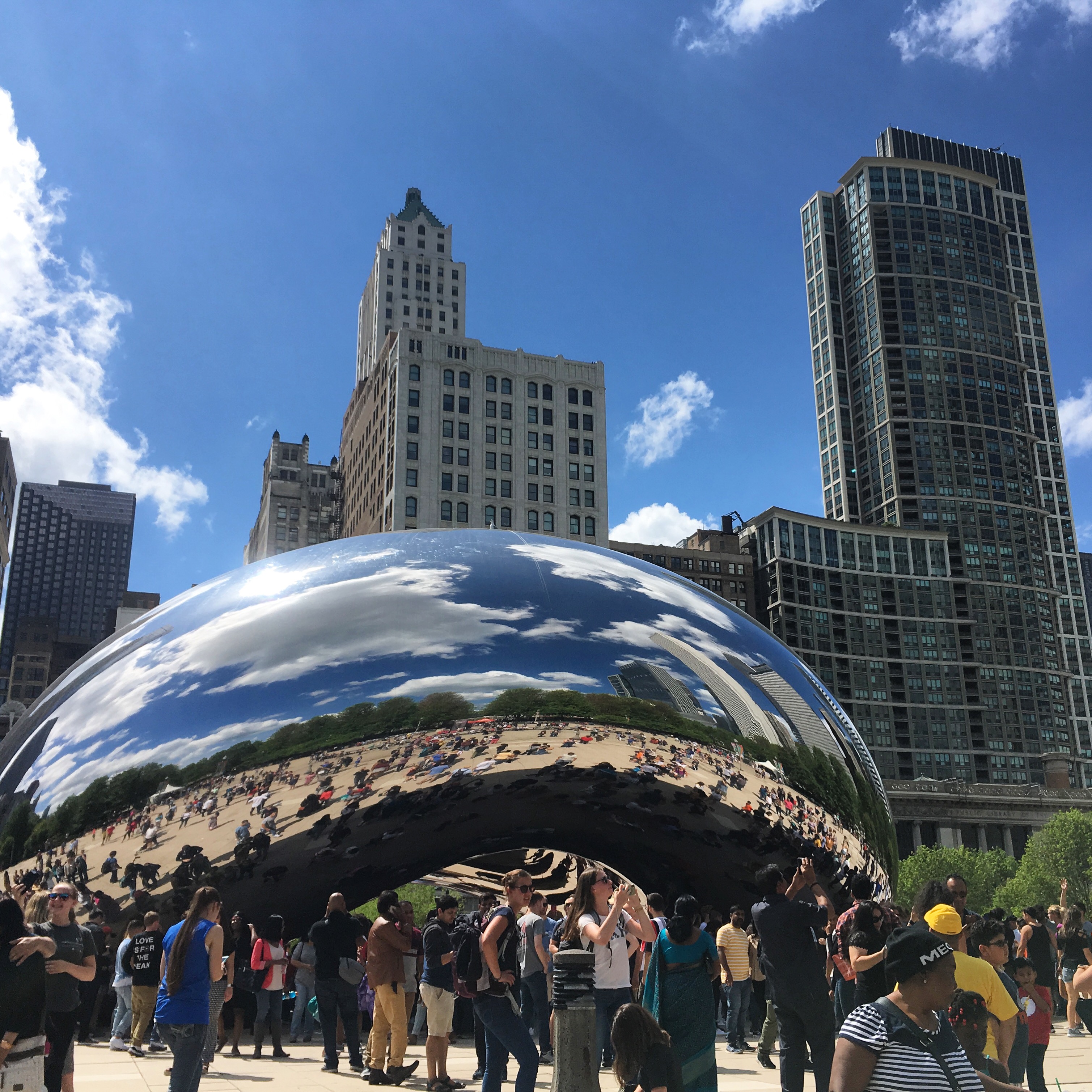 A view of 'the Bean,' formally known as Cloud Gate, a mirrored sculpture in the shape of a bean in Chicago's Millennium Park. In the sculpture you can see buildings reflections, and also behind it are more buildings. There is a crowd of people in front of the sculpture.