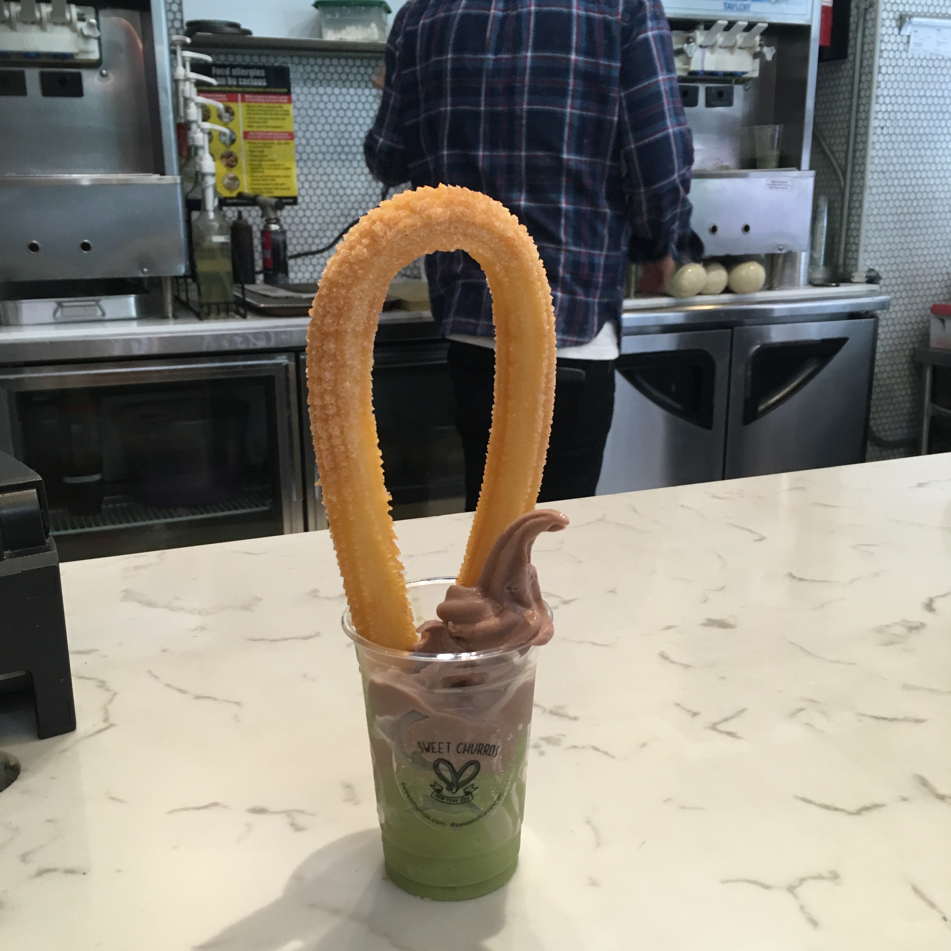 A plastic cup on a counter filled with green matcha ice cream, chocolate ice cream, and a large churro loop.