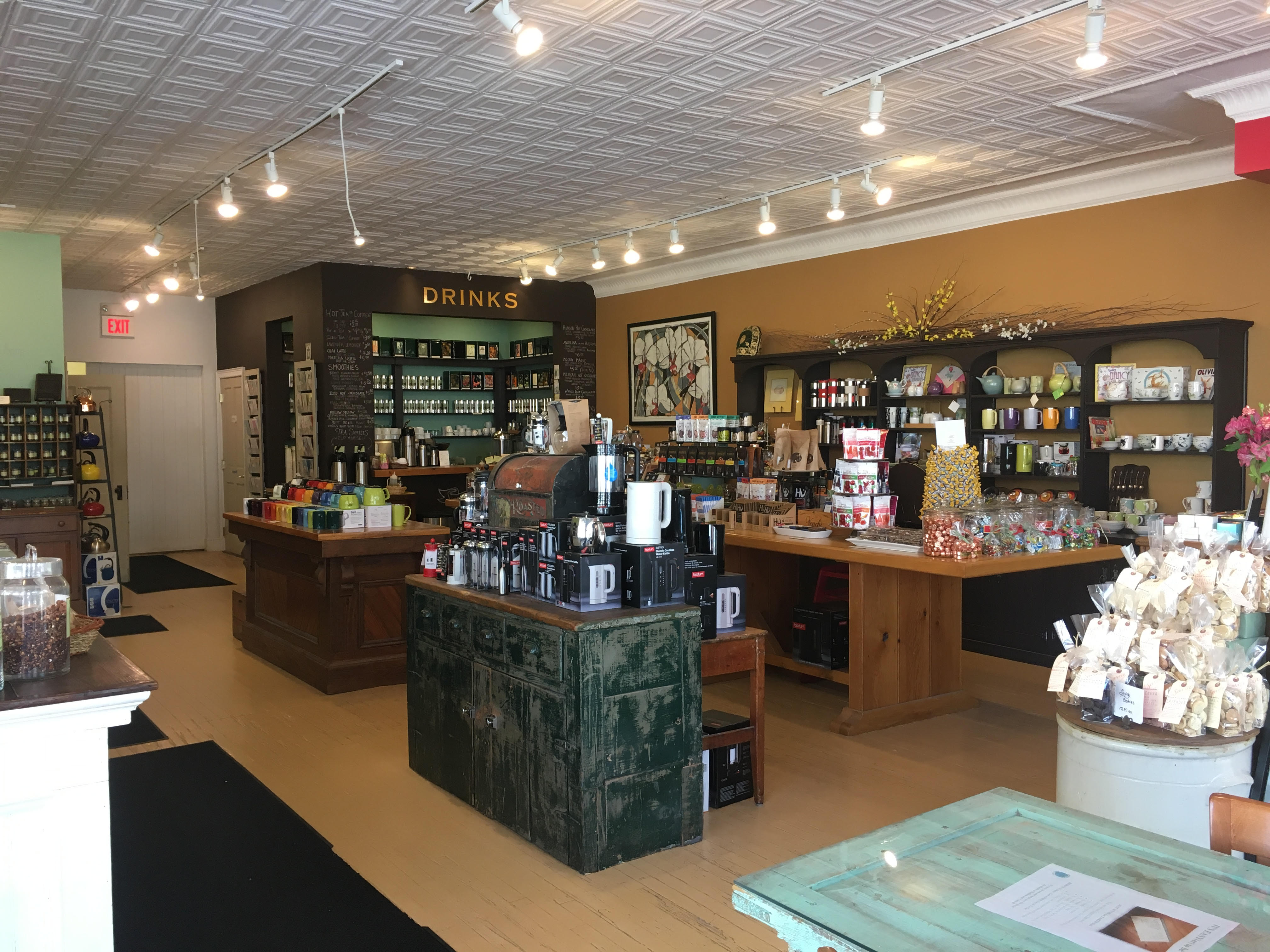 A look into Verdigris shop. There are tables and counters with different items for sale such as teacups, mugs, chocolates in bags. In the back there is another counter with a sign that says "Drinks" over it.